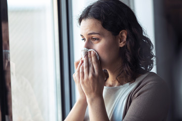 Sad young woman crying while looking through the window at home.