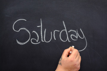 Woman's hand writing the day of the week on a blackboard with white chalk, saturday