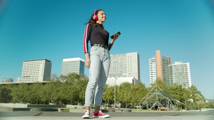 Empowered Young Latin Woman plays music with the mobile using wireless red headphones and starts dancing on a sunny day at the city
