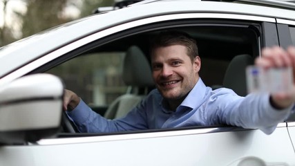 young man sitting in the car and showing his new driver license with thumb up sign out of car window