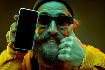 Caucasian woman's portrait on gradient background in neon light. Beautiful male model with hipster style in glasses. Concept of human emotions, facial expression, sales, ad. Showing blank screen phone