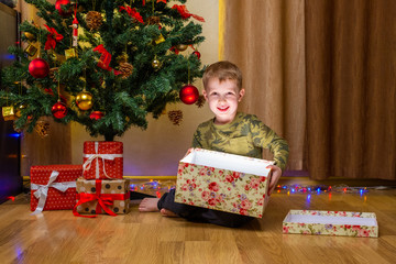 Obraz na płótnie Canvas Happy child boy opening gift box near decorated fir tree. Merry Christmas celebration at home. Little smiling kid playing, having fun with present box with magic light. New Year family holidays.