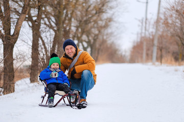 Happy family with kid sledding, having fun and playing with snow outdoors in winter nature. Man, father and child boy on Christmas holidays walking in a snowy park. Wintertime vacation.