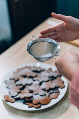 Woman Hand Sprinkling Gingerbread Men with Powdered Sugar. Gingerbread on a Tray in a Wooden table. Christmas cookies homemade.