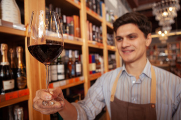 Selective focus on a red wine glass cheerful sommelier is holding out. Happy winemaker enjoying working at his store