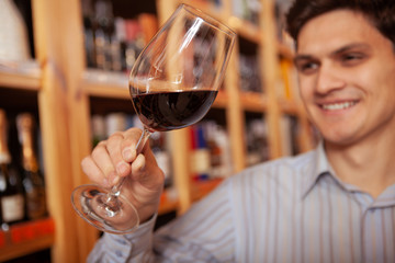 Close up of a red wine glass in the hand of a cheerful man at wine store. Happy male customer tasting red wine prior to buying