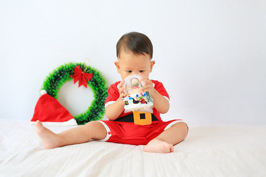 Little Asian baby boy in Santa costume with holding snow globe sitting on bed over white background. Merry Christmas and Happy New Year Concept. - Powered by Adobe