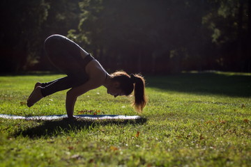 beautiful young girl in a gray jumpsuit practices yoga in a green park in the summer and demonstrates a crane pose or bakasana
