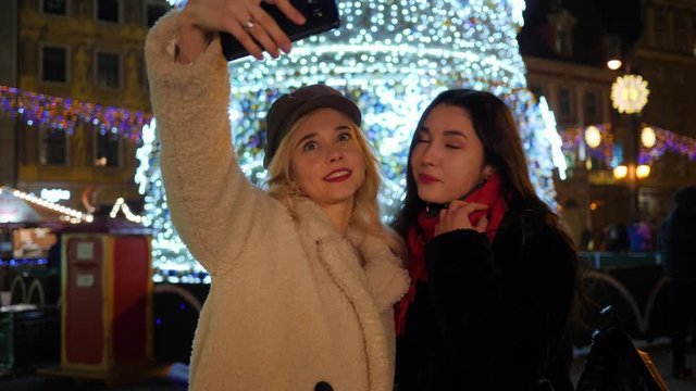 Two pretty young girlfriends women take selfie picture near christmas tree in night city center in Poland
