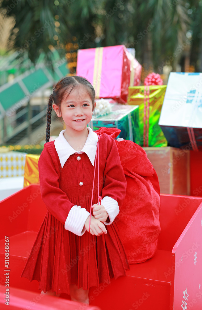 Wall mural Cheerful Asian little girl in red dress carrying a red big bag in winter season and happy new year festival against christmas background.