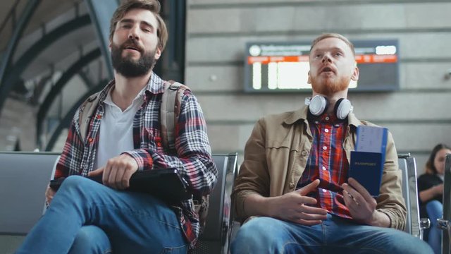 Portrait of young hipsters with luggage sitting down on bench waiting for flight in airport