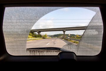 dirty car view from inside the car through the rear window, back window of a dirty car with a view of the asphalt road with markings and a road bridge. - Powered by Adobe