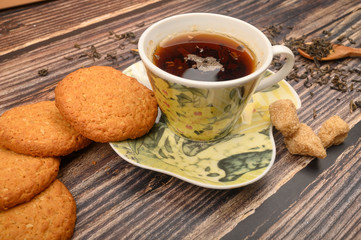 A Cup of black tea, tea leaves, pieces of brown sugar, oatmeal cookies on a wooden background. Close up.
