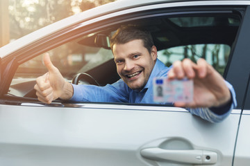 young smiling man sitting in the car and showing his new driver license with thumb up sign out of...