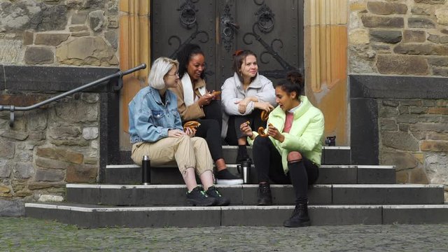 Four Multiracial Smiling Female Teens Eating Pretzels and Drinking Coffee or Tea on the Steps of Ancient European Building. Friendship, Lifestyle and People Concept