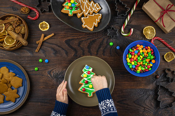 The child decorates with candys a gingerbread in the shape of a Christmas tree. Flat lay with children's hands, lots of sweets on a wooden table.