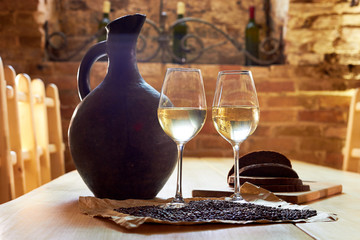 Two glasses of white wine, grape seeds, a clay jug and a few pieces of bread on a wooden table in a restaurant