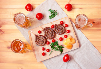 Beer mugs and cutting board with roasted sausages, cherry tomatoes, yellow pepper and fresh parsley on wooden table.