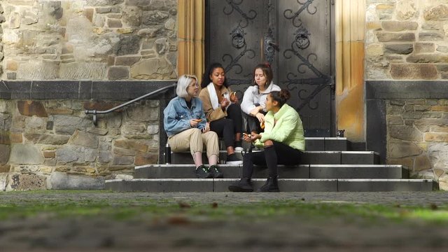 Four Mixed Race Young Girlfriends Eating Pretzels and Talking while Sitting on the Steps of Ancient European Building. Slow Motion. Street Food and Fast food Concept