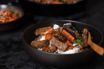 Vegan Korean seitan, carrot and eggplant fry over rice with kimchi in a black bowl