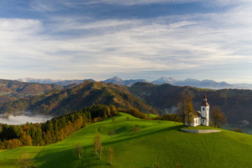 Aerial view of the hills, colorful forest in the fog and the church of Sv Tomaz. Sunrise in...