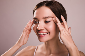 Fototapeta premium Happy positive young woman look to left and smile. Hold hands aside of head. Cheerful positive model touching skin close to eyes. Isolated over beige background. Wrinkles concept. Anti-age