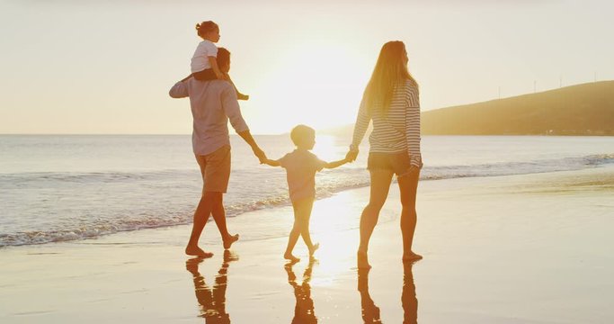 Happy family walking on the beach together, two young boys with their parents, taking a ride on dad's shoulders, holding mom and dad's hands at sunset, happy family on vacation together
