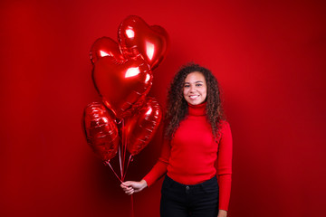 Obraz na płótnie Canvas Studio portrait of young woman with dark skin and long curly hair wearing knitted turtle neck sweater over the festive red wall with heart shaped balloon. Close up, isolated background, copy space.