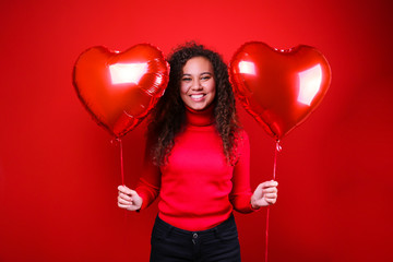 Studio portrait of young woman with dark skin and long curly hair wearing knitted turtle neck sweater over the festive red wall with heart shaped balloon. Close up, isolated background, copy space.