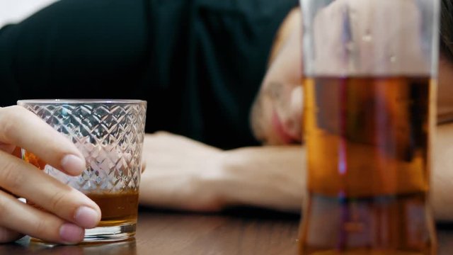 In the background a blurred young drunk man drinks whiskey from a glass and falls asleep on the table, a half-empty bottle of whiskey in the foreground