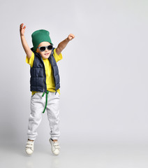Stylish cheerful little boy 4 years old in sweatpants, a green hat, glasses and a down vest has fun