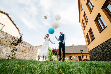 Obraz premium Stylish happy bride and groom hold balloons in hands and jump, poses. On the background on the nature in the courtyard of the house. stone wall. The wind blows the balls.