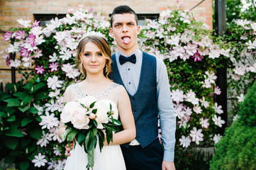 Stylish happy bride with bouquet of peonies with a crown and groom. Husband hugs woman. Close up. Clematis. flowers are gently pink and purple and green leaves. bushes, trees. The groom shows tongue.