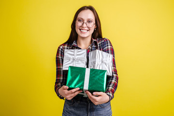 Happy beautiful girl in plaid shirt holding gift boxes enjoying gifts standing on yellow background