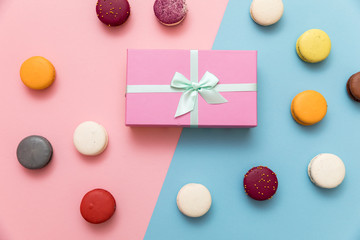 Multi-colored macaroon cakes on a pink and blue pastel background with a gift box flat lay