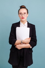 Woman in office suit with glasses holding clean white sheets of paper standing on blue background...