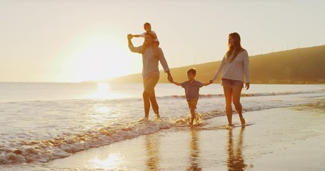 Happy family walking on the beach together at sunset, two young boys and their parents on the beach, riding on dad's shoulders, holding hands with mom and dad