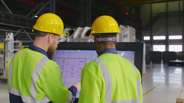 Two diverse male engineers in uniform and hard hats use computer LCD touch screen for programming CNC machine. Work process on heavy industry plant or factory. Employees and high tech