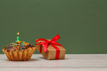 Birthday cupcake with burning festive candle and gift box on green background.