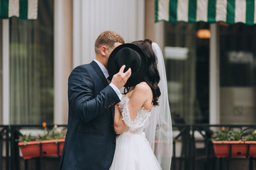 The groom in a blue suit and the bride in a white dress kiss and hide behind a black hat. Photography, concept. Wedding portrait.