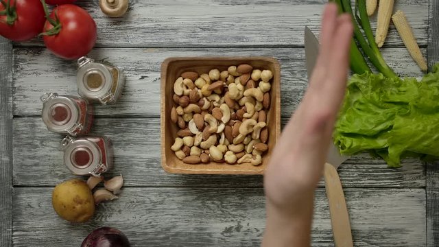 The hand pours the nuts into a wooden bowl on the kitchen table. Spices, potatoes, tomatoes, lettuce, pepper and corn are all around.