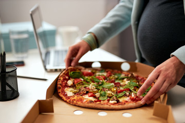 Close up of caucasian pregnant business woman in forties and dressed in suit standing in office and having pizza for lunch.