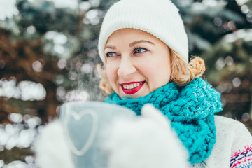 Happy girl with a cup of hot coffee in the cold - in white warm mittens, a scarf and a hat with a cup of hot drink in the forest - a nice New Year and Christmas look