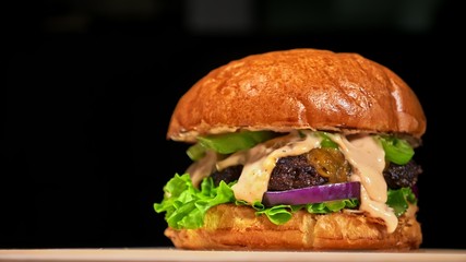 Craft burger is cooking on black background. Consist: red sauce salsa, lettuce, red onion, pickle, cheese, chilli green pepper, air bun and marbled meat beef. Not made ideal. Loving hand made.