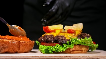 Craft burger is cooking on black background in black food gloves. Consist: red sauce salsa, lettuce, red onion, pickle, cheese, chilli green pepper, air bun and marbled meat beef. Not made ideal