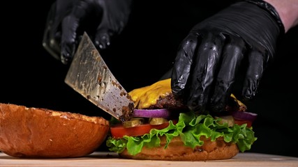 Craft burger is cooking on black background in black food gloves. Consist: sauce, lettuce, tomato, red onion, pickle, cheese, bacon, air bun and marbled meat beef. Not made ideal. Looks real, loving
