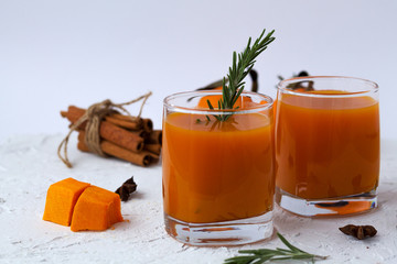 two glasses of fresh pumpkin juice and oranges with slices of ripe pumpkin and spices on a white background. fresh cocktail with sprig of rosemary and sticks of cinnamon with mandarins on white table.