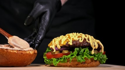 Craft burger is cooking on black background in black food gloves. Consist: sauce, lettuce, tomato, red onion, pickle, cheese, bacon, air bun and marbled meat beef. Not made ideal. Looks real, loving