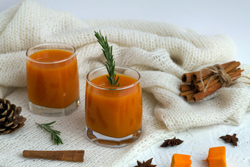cocktail with sprig of rosemary and sticks of cinnamon on the warm white plaid on white table. two glasses of fresh pumpkin or oranges juice with slices of pumpkin, spices and winter decoration.