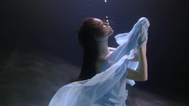 a beautiful girl with long hair and a sequined dress is floating under the water. she turns in profile and holds out her hands with the blue cloth